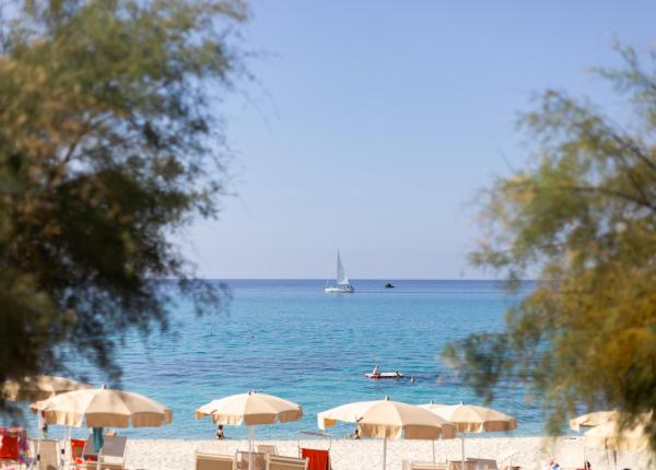 Spiaggia con ombrelloni, mare calmo e barca a vela all'orizzonte.