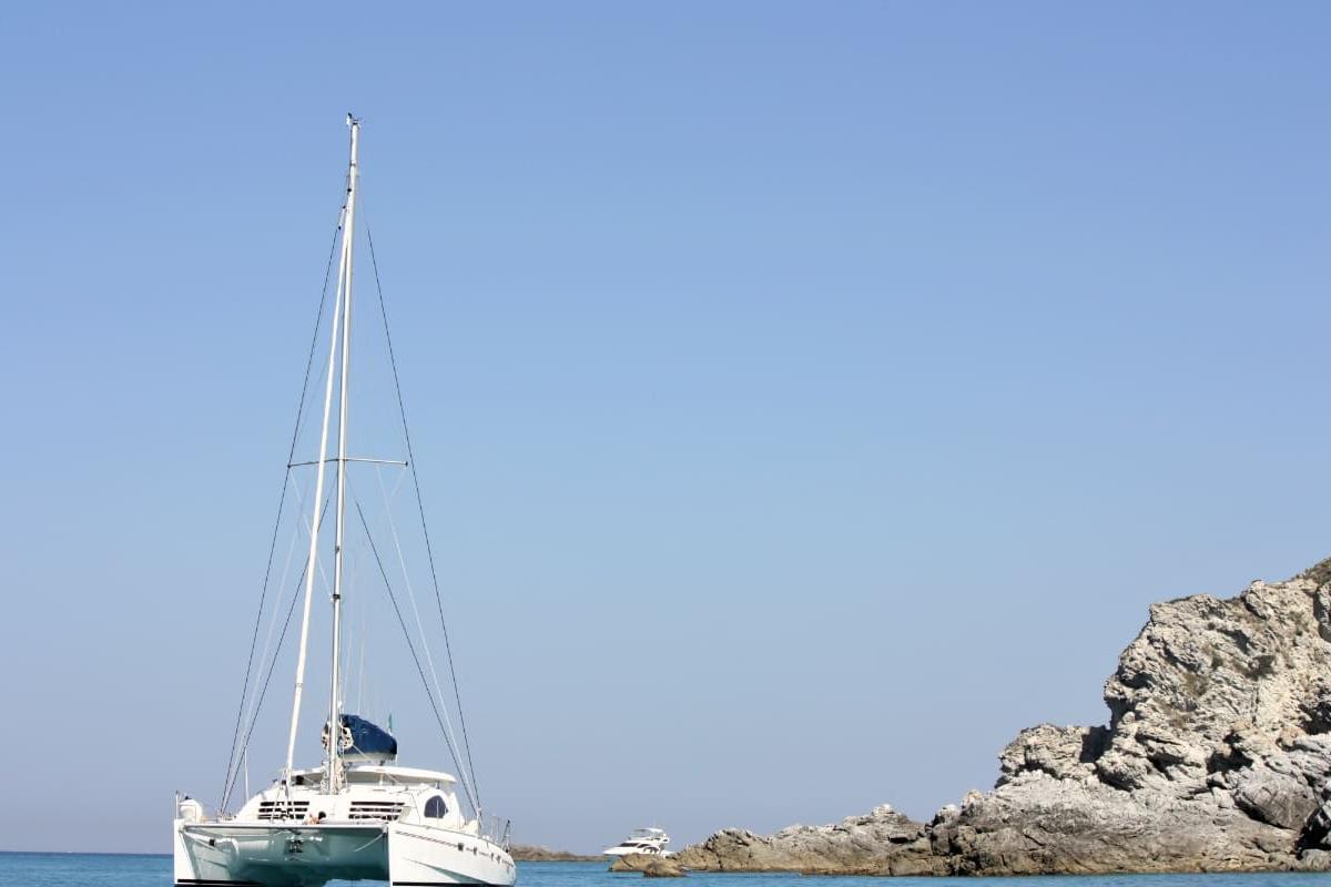 Catamarano bianco vicino a una costa rocciosa sotto un cielo azzurro.