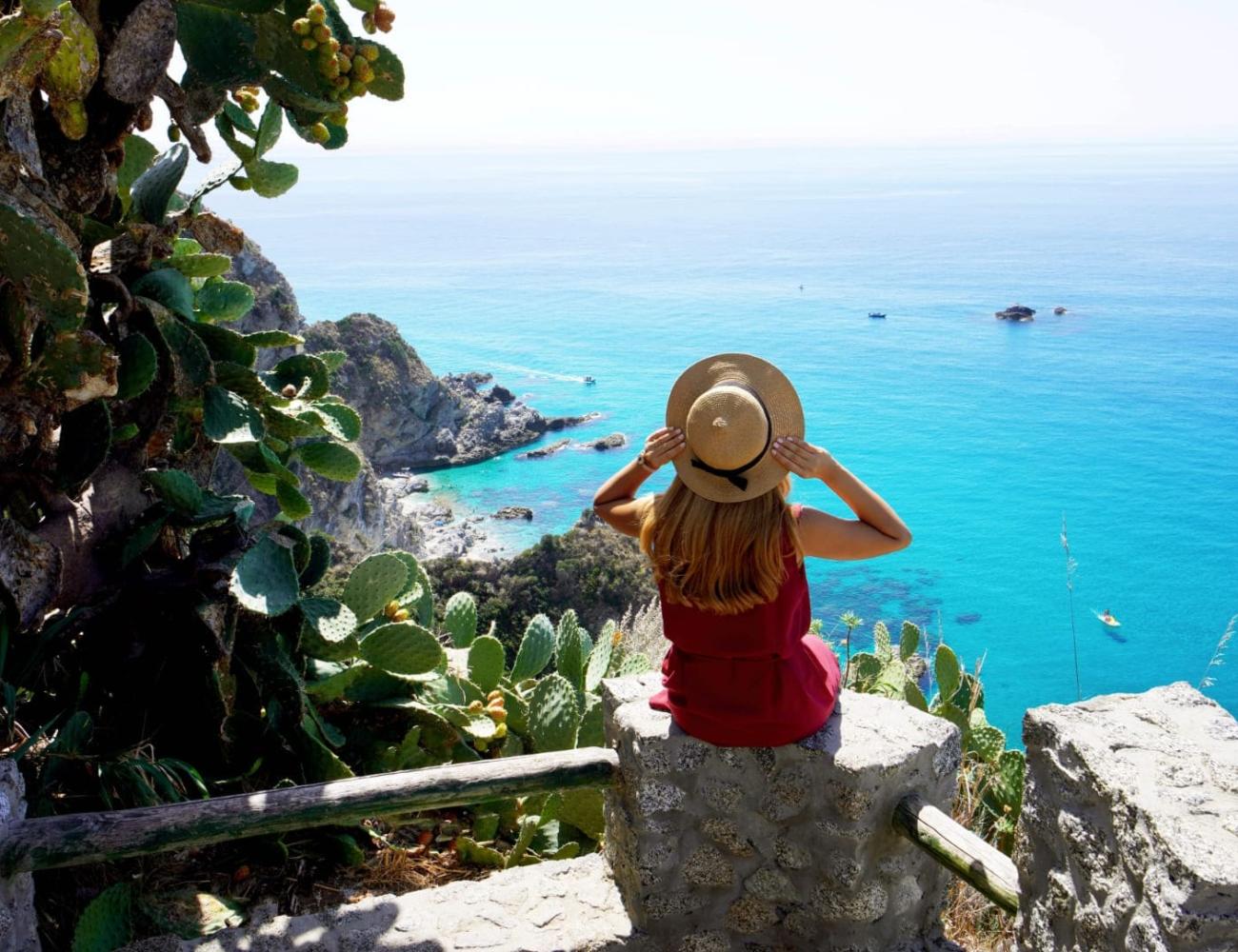 Woman with hat admires turquoise sea from a cliff.