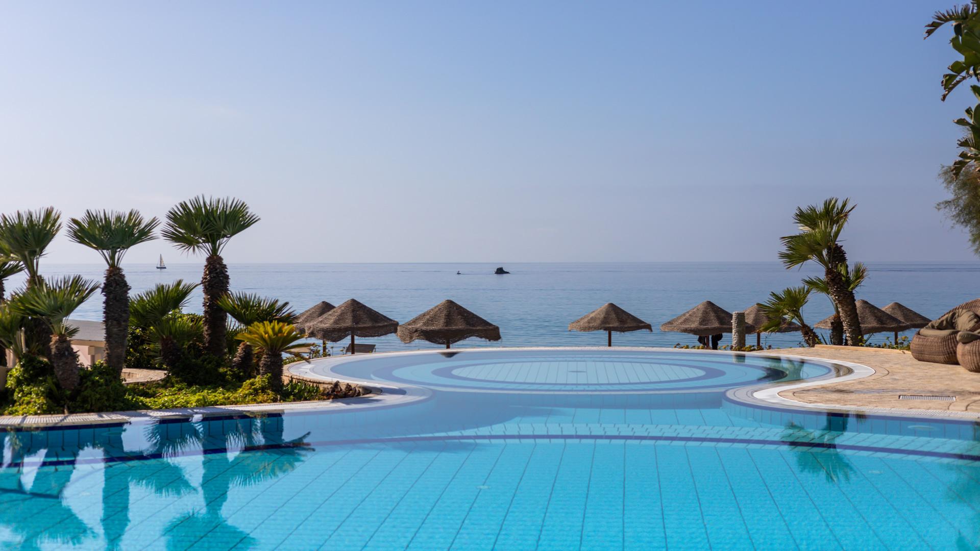 Piscina con vista mare, palme e ombrelloni di paglia.