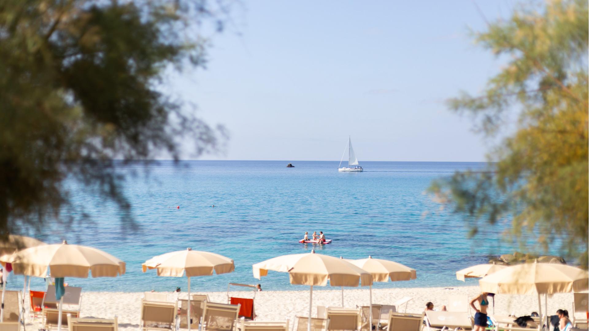 Spiaggia sabbiosa con ombrelloni, mare azzurro e barca a vela all'orizzonte.