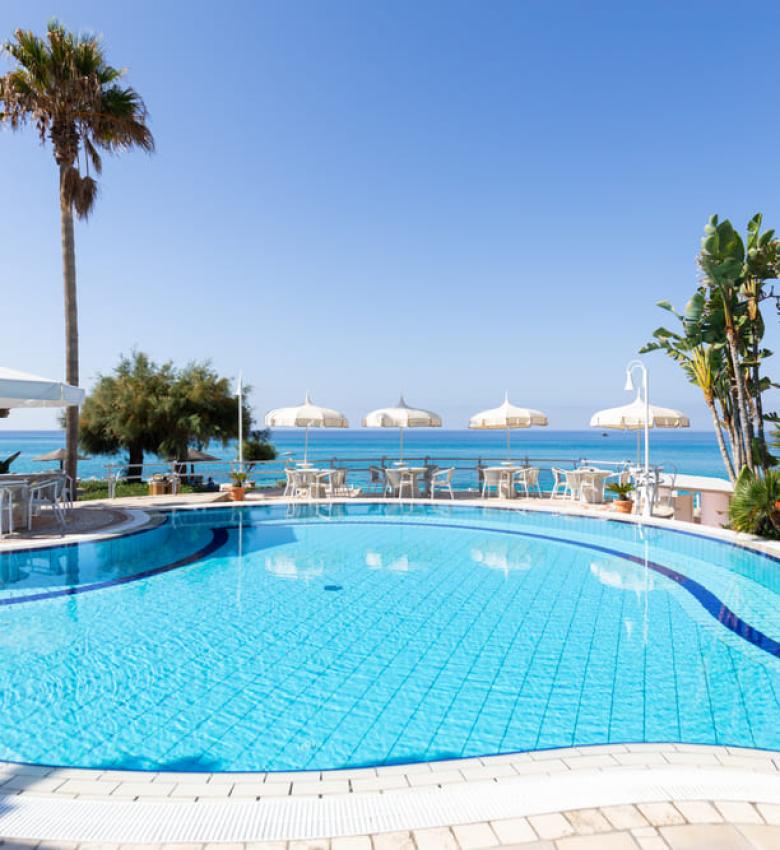 Piscina con vista mare, palme e ombrelloni bianchi.